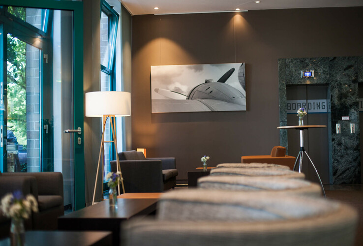 Modern hotel lobby with seating, large lamp and picture of an airplane on the wall at the ATLANTIC Hotel Airport Bremen.