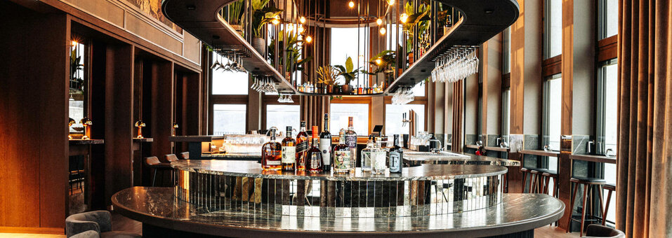 Elegante Hotelbar mit runder Theke, Flaschen, Gläsern und Pflanzen, umgeben von hohen Fenstern und gemütlichen Sitzgelegenheiten.
