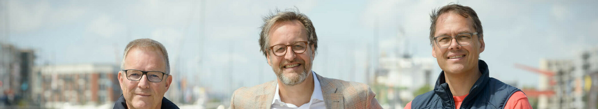 Verbundausbildung im ATLANTIC Hotel Sail City Bremerhaven Drei Männer stehen lächelnd vor einem Geländer mit Wasser und Hafen im Hintergrund.