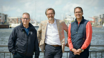 Drei Männer stehen lächelnd vor einem Geländer mit Wasser und Hafen im Hintergrund.