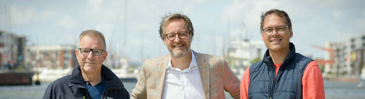 Drei Männer stehen lächelnd vor einem Geländer mit Wasser und Hafen im Hintergrund.