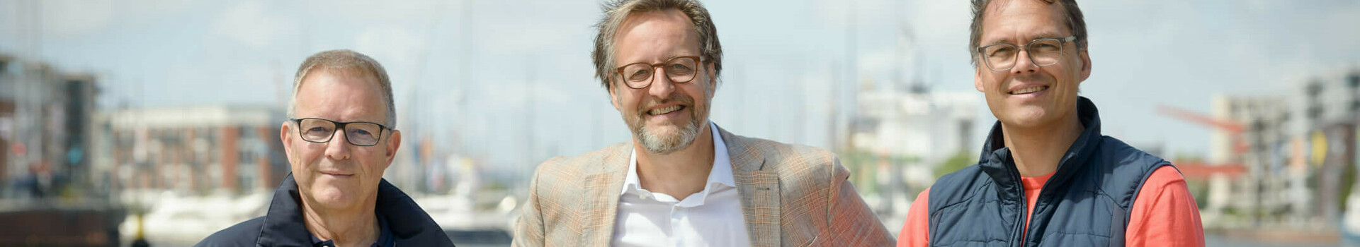 Drei Männer stehen lächelnd vor einem Geländer mit Wasser und Hafen im Hintergrund.