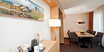 Helles Hotelzimmer mit modernem Holzmobiliar, Sitzecke, Tisch, Kaffeestation und großem Landschaftsbild an der Wand.