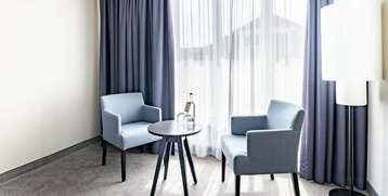 Helles Hotelzimmer mit zwei blauen Sesseln, Tisch, Wasserflasche und Stehlampe vor einem großen Fenster mit Vorhängen.