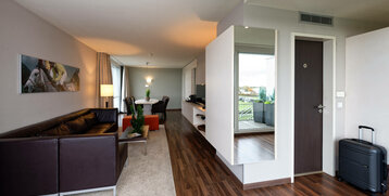 Modern hotel room with leather sofa, dining table, artwork on the wall and view of balcony with plants.
