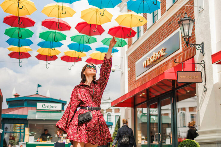 Frau in rotem Kleid spaziert unter bunten, schwebenden Regenschirmen in einer Einkaufsstraße.
