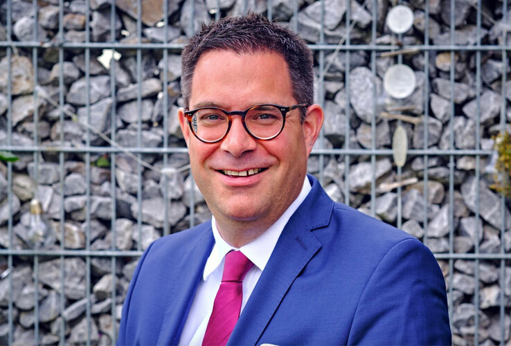 Ein lächelnder Mann in Anzug vor einer Steinmauer.