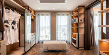 Heller, moderner Ankleidebereich im ATLANTIC Hotel Heidelberg mit Kleiderschrank, Spiegel, Sitzbank und großen Fenstern.