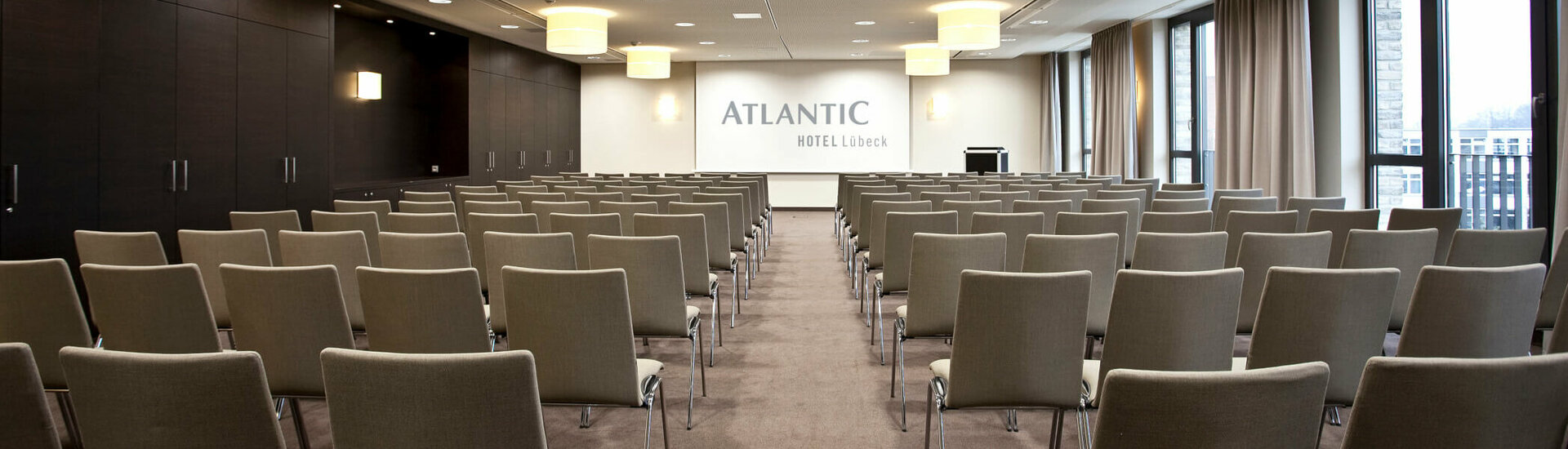Modern conference room in the ATLANTIC Hotel Lübeck with rows of chairs, stage and large windows.