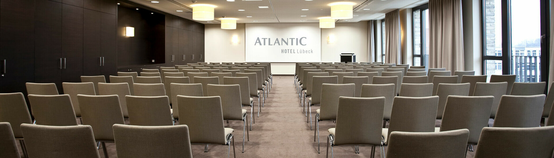 Moderner Konferenzraum im ATLANTIC Hotel Lübeck mit Reihen von Stühlen, Bühne und großen Fenstern.