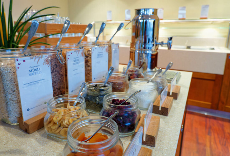 Müsli-Buffet mit verschiedenen Zutaten in Gläsern und einem großen Getränkespender im Hintergrund.