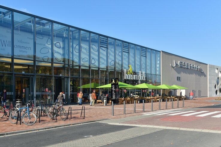 Einkaufszentrum mit Glasfassade, Restaurantterrasse mit grünen Schirmen, Fahrradständer und Menschen im Eingangsbereich.