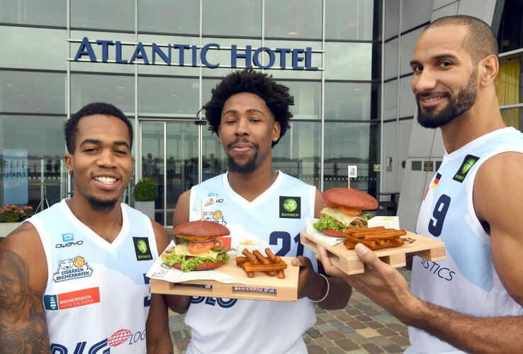 Three basketball players in front of the ATLANTIC Hotel with burgers and fries in their hands, smiling.