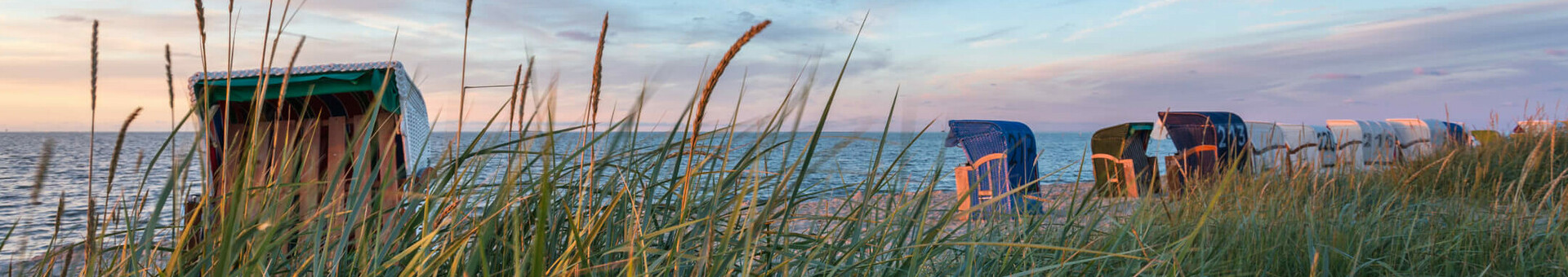 Angebote und Arrangements der ATLANTIC Hotels Strandkörbe am Meer bei Sonnenuntergang, umgeben von Dünen und hohem Gras, blauer Himmel im Hintergrund.