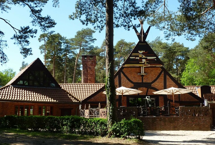 Backsteingebäude im Wald mit Terrassenbereich, Sonnenschirmen und dekorativen Holzelementen unter blauem Himmel.