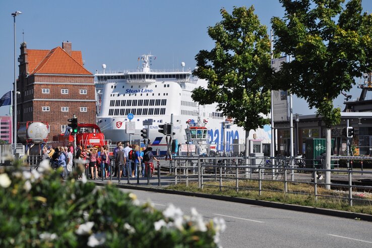 Gruppe von Menschen vor einem großen Fährschiff im Hafen, umgeben von Bäumen und einem Backsteingebäude.