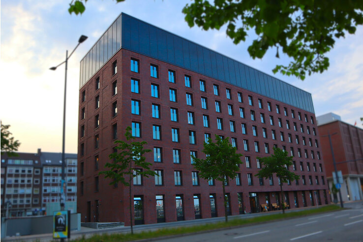 Modern red-brick hotel building with large windows, surrounded by trees and the street in the foreground.