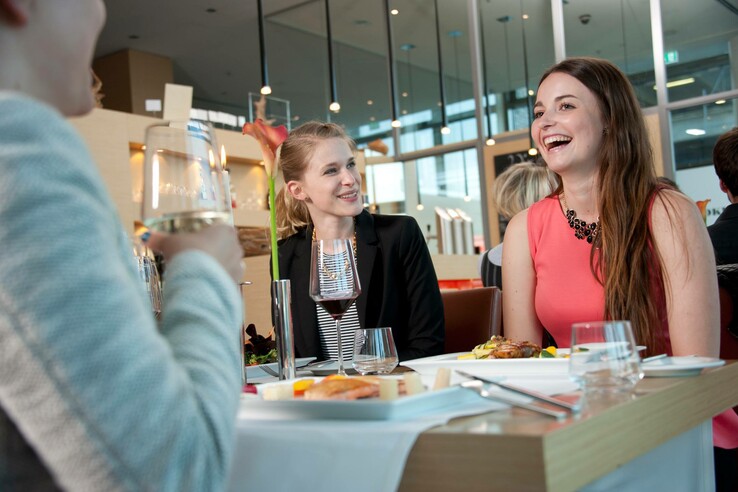 Drei Personen lachen und unterhalten sich beim Essen in einem modernen Restaurant des ATLANTIC Hotels Kiel.
