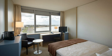 Bright hotel room with double bed, desk, TV and panoramic window overlooking the river in Bremerhaven.