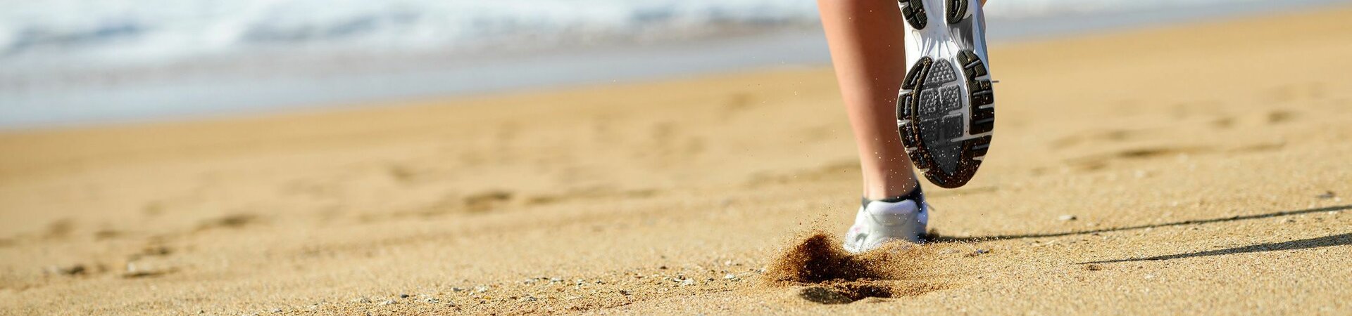 Person joggt am Sandstrand entlang, hinterlässt Fußabdrücke, mit Meer im Hintergrund.