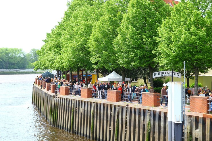 View of a lively beer garden on the riverbank with green trees and people enjoying the view.