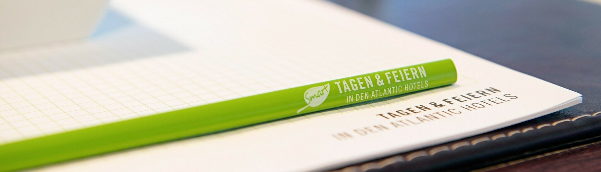 Grüner Stift und Block mit "Tagen & Feiern" Aufschrift auf einem Konferenztisch, daneben ein Glas Wasser und eine Schale.