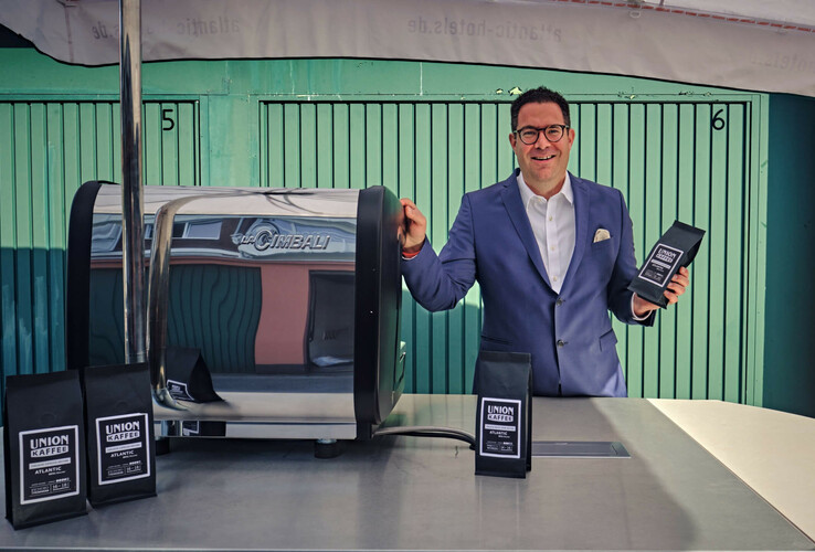 Mann in Anzug präsentiert Kaffeepackung neben Espressomaschine im ATLANTIC Hotel Münster.