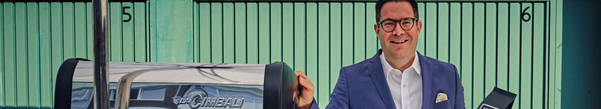 Mann in Anzug präsentiert Kaffeepackung neben Espressomaschine im ATLANTIC Hotel Münster.