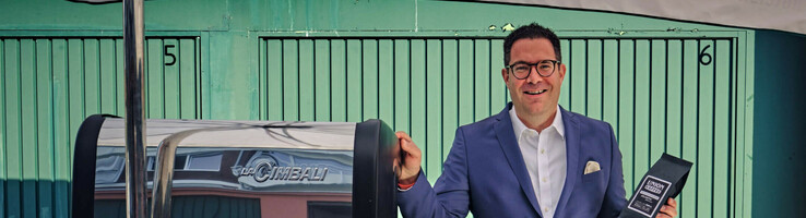 Hoteldirektor mit Kaffeerad – ATLANTIC Hotel Münster Mann in Anzug präsentiert Kaffeepackung neben Espressomaschine im ATLANTIC Hotel Münster.