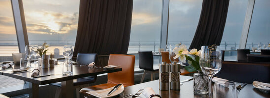 Modernes Restaurant mit gedeckten Tischen und Meerblick bei Sonnenuntergang im ATLANTIC Hotel Sail City, Bremerhaven.