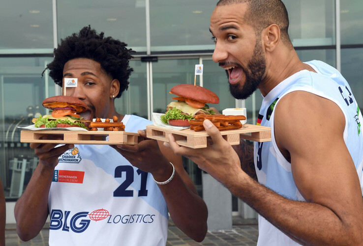 Two men in basketball jerseys eating burgers in front of the ATLANTIC Hotel Sail City in Bremerhaven.