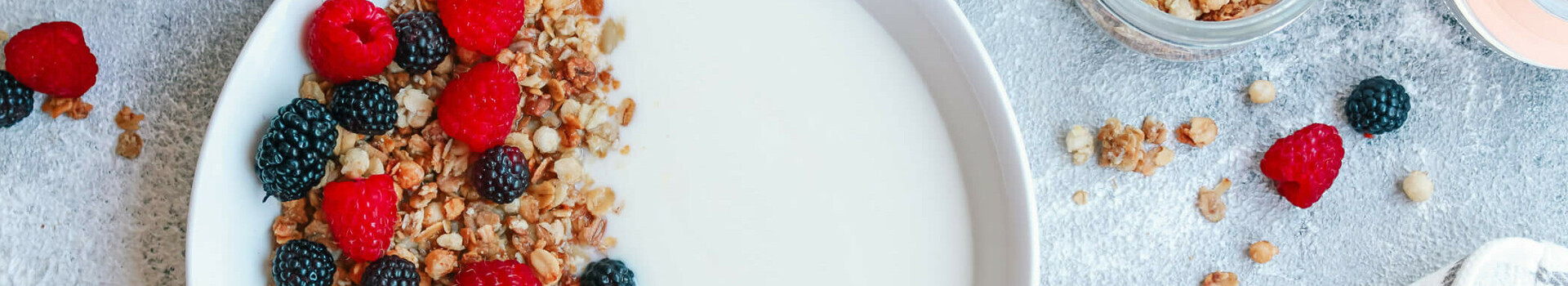 Schale mit Joghurt, Müsli, Himbeeren und Brombeeren, umgeben von gefüllten Gläsern und einem gestreiften Tuch.