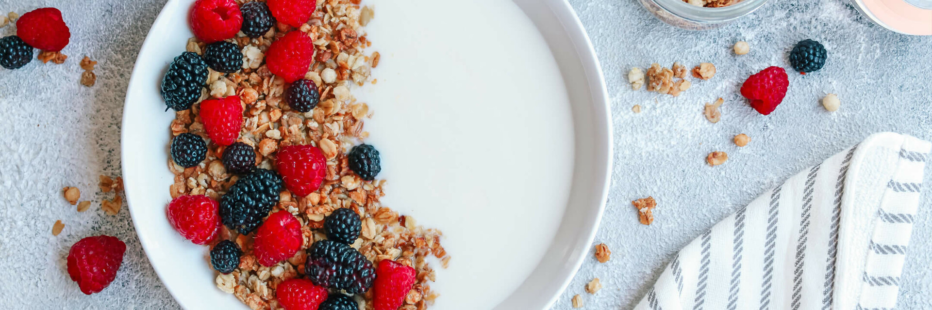 Schale mit Joghurt, Müsli, Himbeeren und Brombeeren, umgeben von gefüllten Gläsern und einem gestreiften Tuch.