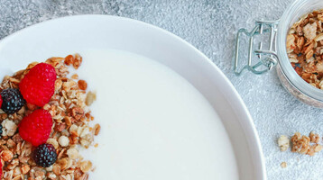 Schale mit Joghurt, Müsli, Himbeeren und Brombeeren, umgeben von gefüllten Gläsern und einem gestreiften Tuch.