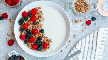 breakfast in the ATLANTIC Hotel Wilhelmshaven Bowl with yogurt, muesli, raspberries and blackberries, surrounded by filled glasses and a striped cloth.