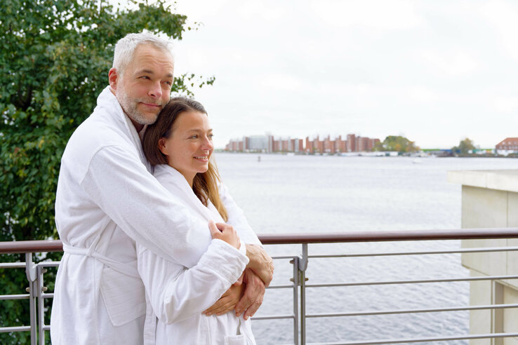 Couple in bathrobes hugging on balcony with lake view.