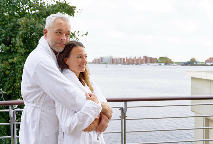 Paar in Bademänteln umarmt sich auf Balkon mit Seeblick.