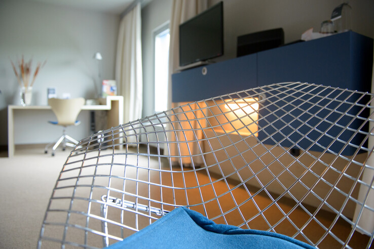 Modernes Hotelzimmer mit Schreibtisch, Stuhl, blauem Sideboard und stilvollem Metallstuhl im Vordergrund.