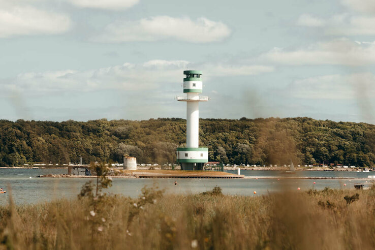Weiß-grüner Leuchtturm am Wasser, umgeben von Wald und Wiese, unter blauem Himmel mit Wolken.