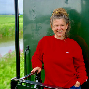 Eine lächelnde Person in rotem Pullover steht draußen vor grüner Landschaft.