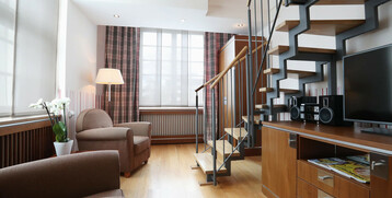 Helles Hotelzimmer mit moderner Treppe, gemütlichen Sesseln, Lampe, TV und Orchidee auf Holzboden.