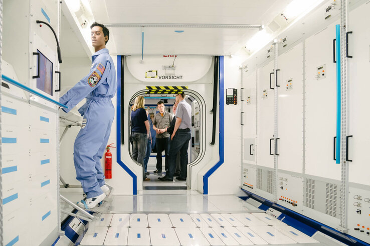 Person in blauem Raumanzug in einem weißen, futuristischen Korridor mit Technik, im Hintergrund stehen mehrere Personen.