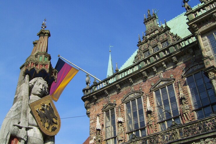 Statue mit Schild und Schwert vor historischem Gebäude, daneben weht eine deutsche Flagge, blauer Himmel im Hintergrund.