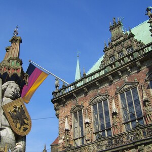 The Bremer Roland on the market place  Statue with shield and sword in front of a historic building, a German flag flying next to it, blue sky in the background.