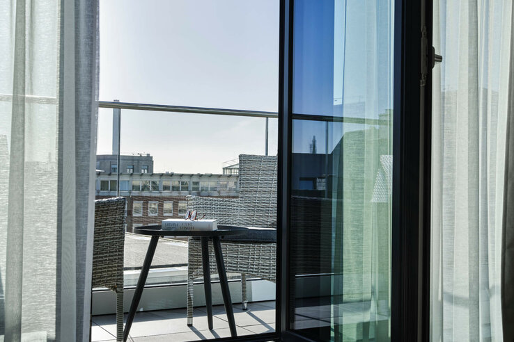 Balcony with table, chair and book, view of city building through glass window, light curtains in the ATLANTIC Hotel Münster.