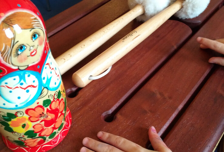 Ein Xylophon mit Schlägeln und einer bemalten Matroschka-Puppe auf einem Holztisch. Kinderhände greifen danach.