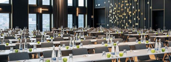 Moderner Konferenzraum im ATLANTIC Hotel Heidelberg mit Reihen von Tischen und Stühlen, großen Fenstern und eleganter Beleuchtung.