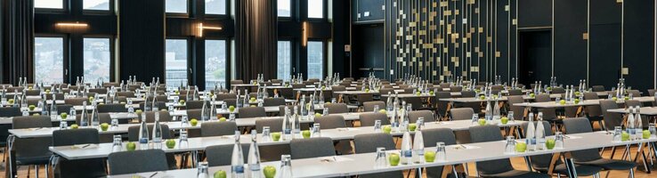 ATLANTIC Hotel Heidelberg Ballsaal mit parlamentarischer Bestuhlung Moderner Konferenzraum im ATLANTIC Hotel Heidelberg mit Reihen von Tischen und Stühlen, großen Fenstern und eleganter Beleuchtung.