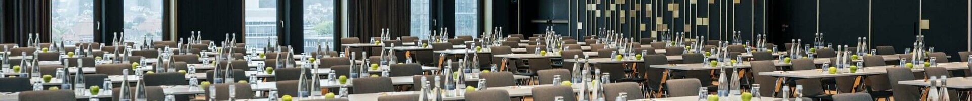 Moderner Konferenzraum im ATLANTIC Hotel Heidelberg mit Reihen von Tischen und Stühlen, großen Fenstern und eleganter Beleuchtung.