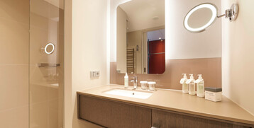 Modernes Badezimmer im ATLANTIC Grand Hotel Bremen mit beleuchtetem Spiegel, Waschbecken und Pflegeprodukten auf der Ablage.